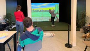 A person tries out a golf simulator while three other people watch. 