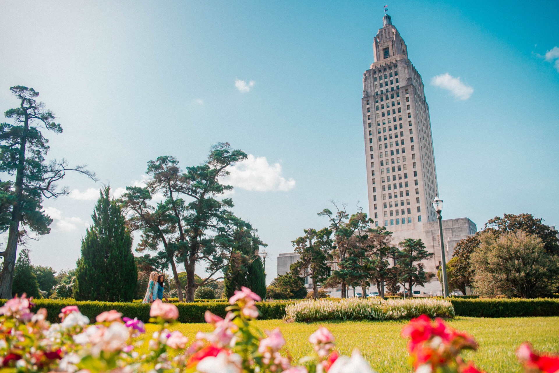 The Louisiana state capitol building towers over a vibrant park filled with blooming flowers and lush greenery, under a clear blue sky.