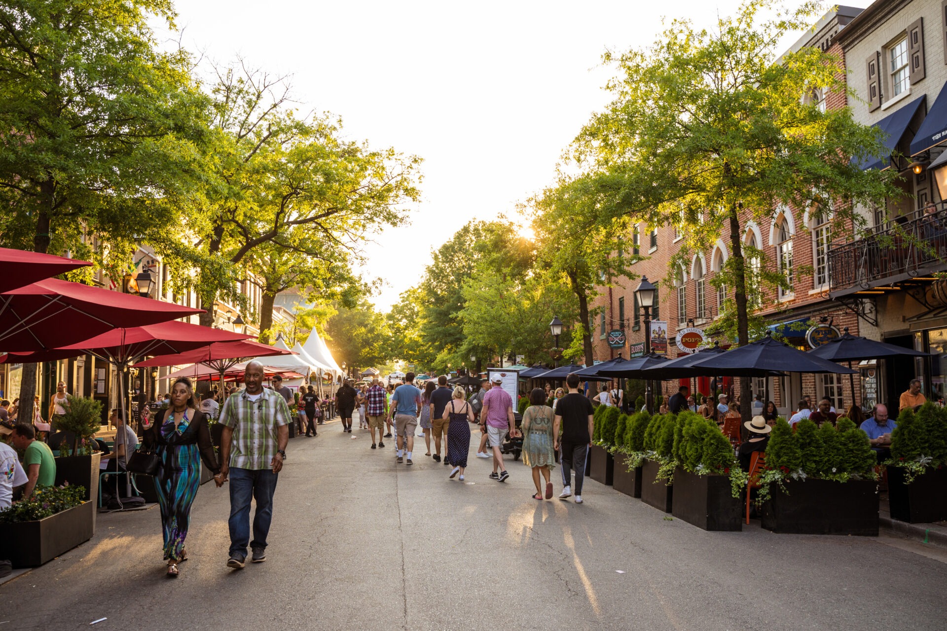 A street with patios and people walking. There are a row of trees on either side of the street.