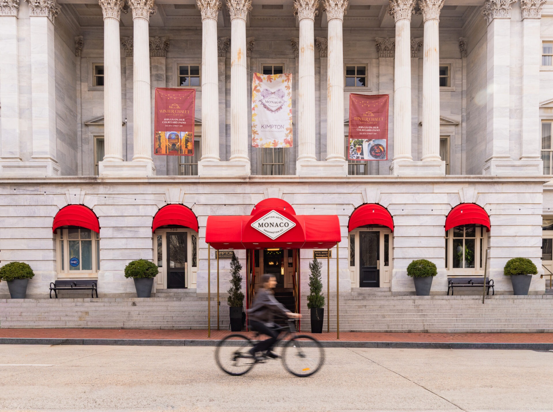 The exterior of a hotel with many columns. Someone is riding a bike in front of it.